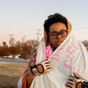 Naima lowe standing in the foreground with a brown headscarf over her naturally standing hair. She is wearing black glasses with her mouth slightly open. Around her neck a white scarf is held in place , she is wearing reflective gold fingerless gloves. The white scarf has pink cursive handwriting printed across it that reads "Fuck the Police" the "i" in police is dotted with a heart. In the background is a street with parked cars and trees with red leaves.