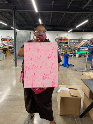 Naima Lowe with braided hair and clear glasses, standing in the foreground holding a light pink print bearing the phrase "Fuck the Police" in darker pink cursive handwriting many times over in different sizes. In the background is a printing studio. 