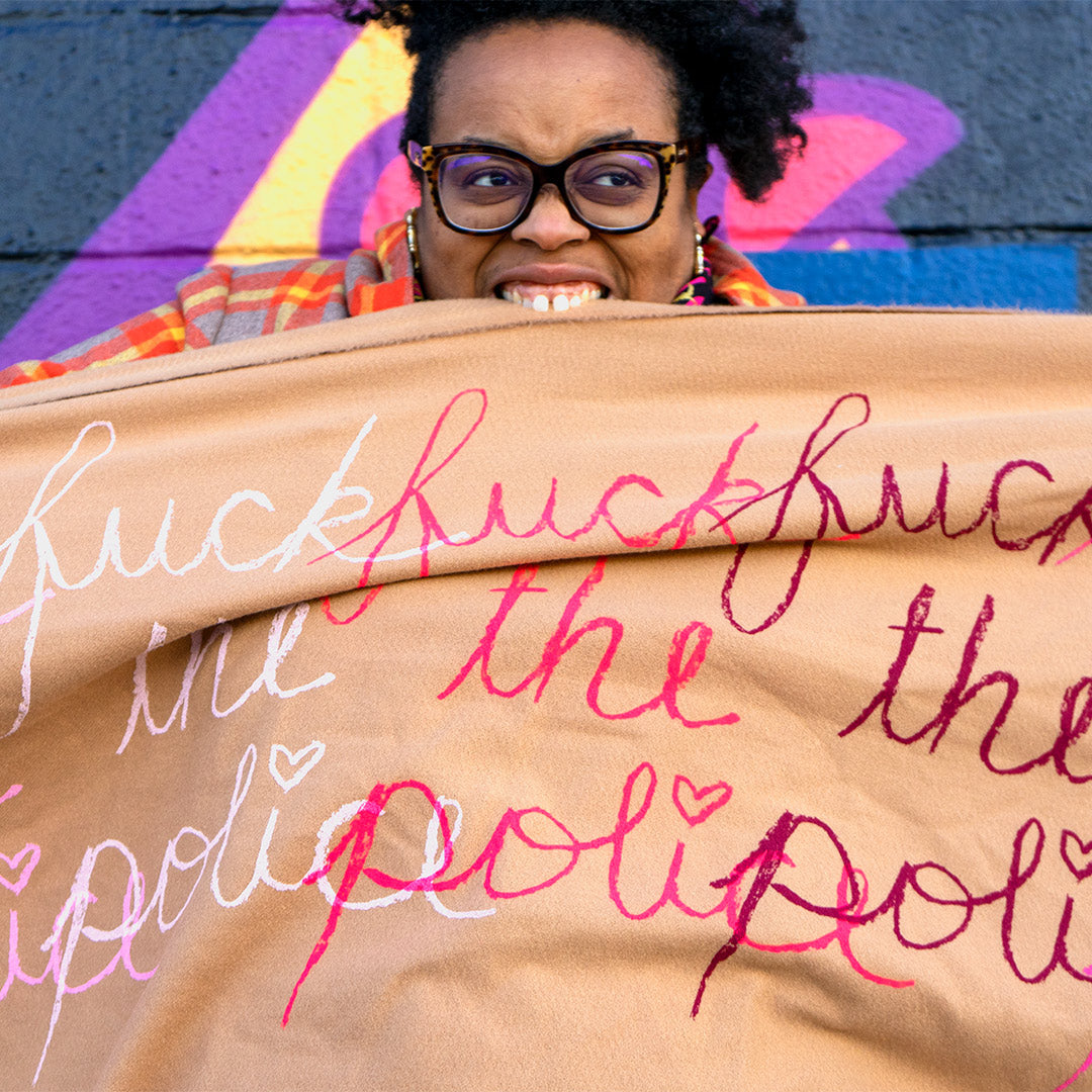 Naima Lowe, standing in front of a black wall with yellow, purple and red writing. Her hair stands up past the frame, she wears large black glasses, and a black, red and yellow dress. She is biting down on a large Khaki-colored scarf, stretched out to fill the frame. Printed on the scarf is cursive handwriting bearing the phrase "fuck the police" 3 times in different colors: white, pink and magenta. The "i" is dotted with a heart, the phrases overlap each other slightly.