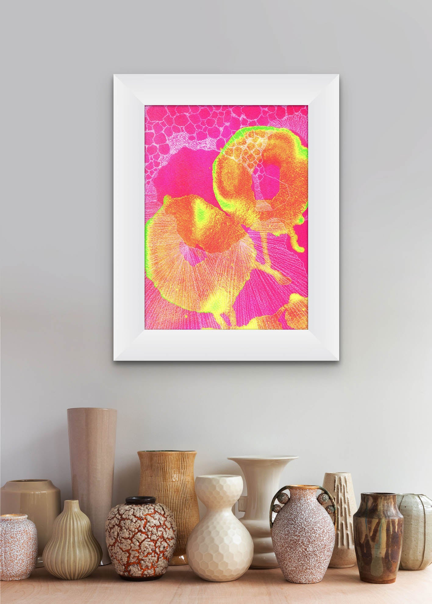 Bright pink, yellow, green and white screen print shown in a white frame in a neutral home space background. 