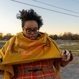 Naima Lowe standing in the foreground under a power-line. Her hair is standing naturally. She is looking down at a large mustard-colored Cashmere scarf printed with cursive handwriting bearing the phrase "Fuck the Police" in 3 different colors, white, pink and dark magenta. The "i" in Police is dotted with a heart. She is wearing fingerless reflective gold gloves and stretching out two sections of the scarf so that the bottom edge of it extends in a straight line. The sky behind her is either dusk or dawn. 