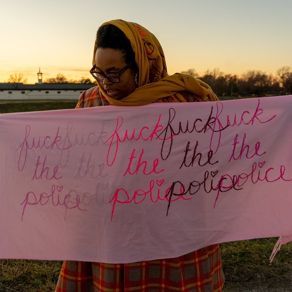 Naima Lowe, wearing a yellow headscarf, black glasses and a black, red and yellow plaid dress, stands in a large clearing holding a light pink scarf which is decorated with the phrase "fuck the police" 5 times, overlapping in different shades of pink