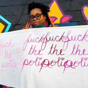 Naima Lowe standing in front of a wall painted with yellow, purple, turquoise, and pink paint. Her hair stands naturally, she is wearing black glasses. A pink, black and turquoise face mask is around her neck. She has a black, pink and red hood stretched around the back of her head. Extended across the bottom half of the frame is a large White scarf with large cursive handwriting bearing the phrase "Fuck the Police," 5 times in 5 different shades of pink, overlapping. 