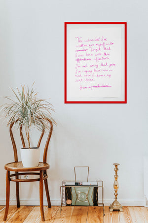 The art print in a thin red frame hanging on the upper right side of cream colored wall. A brass candlestick, bronze magazine holder with a black and light green magazine inside sit on a hardwood floor below the frame. The wall has simple molding at the edges. Next to the magazine holder is a small tropical plant sitting on a polished wooden chair. 